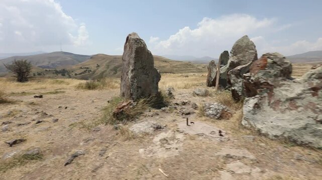 Camera pans across ancient standing stones in a mountainous landscape, revealing a close-up of a distinctive circular hole bored through a rough megalith at Zorats Karer, Armenia.