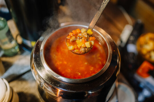 Bowl of hearty vegetable soup being served from a stainless steel pot with steam rising in a rustic kitchen setting