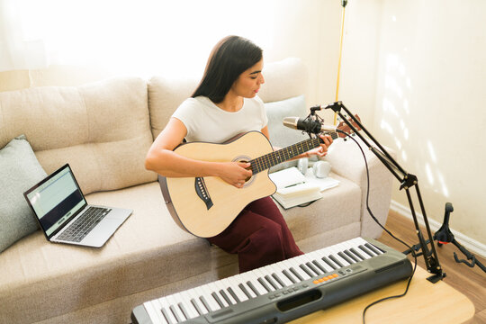 Latin woman composing music at home, playing acoustic guitar and recording song vocals with a microphone