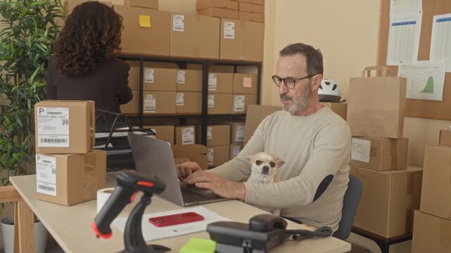 Man typing on laptop with small dog on lap and stacked shipping parcels, scanning labels with barcode reader in building; focused productivity.