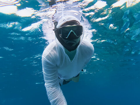 Man in protective white hooded rash guard shirt snorkeling in Red Sea, Egypt, avoiding sunburn under harsh sun while exploring underwater