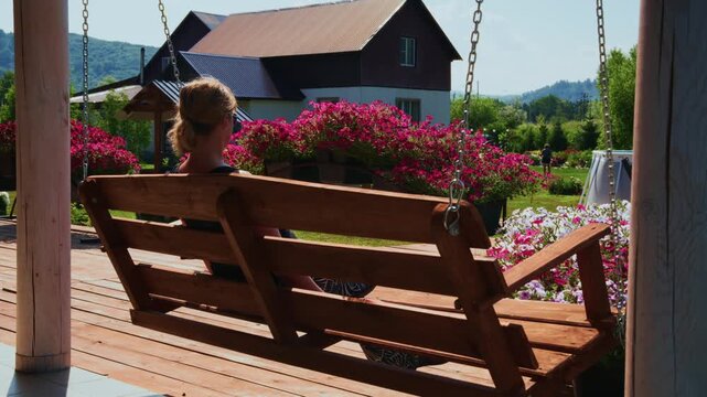 Woman sits calmly on a wooden swing. Her ponytail rests against the back. She gazes toward pink blossoms. House with a brown roof appears. Mountain slopes stretch across the horizon
