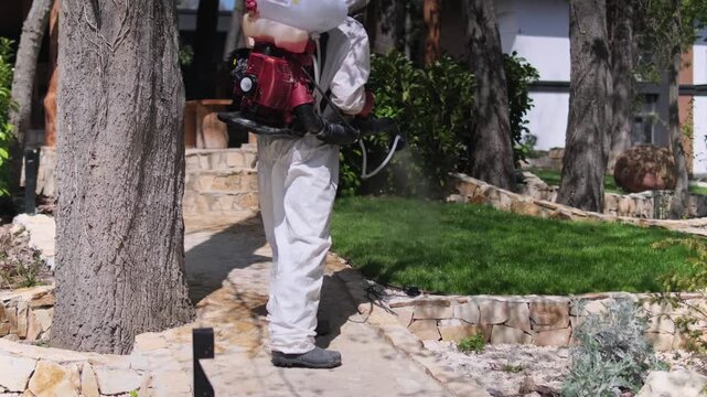 Worker in protective suit spraying garden with pesticide using backpack fogger, outdoor pest control treatment in residential yard