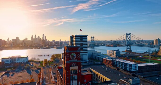 Flying along the Nipper Building located in Camden, New Jersey. The Delaware River with the Benjamin Franklin Bridge across it and skyline of Philadelphia, Pennsylvania, USA at sunset in the backdrop.