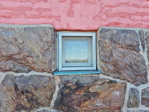 Close-up architectural detail of a small basement window framed by rough natural stone foundation and pink plaster wall. Captured in Sweden.