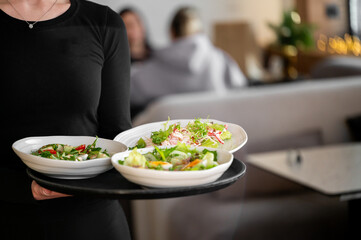 Naklejka premium A waitress in a black uniform holds a black tray with three white bowls of fresh, gourmet green salads in a modern restaurant setting