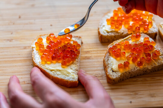 Bread butter sandwich with caviar, woman hand spreading fish roe with spoon macro closeup of wooden cutting board at home kitchen
