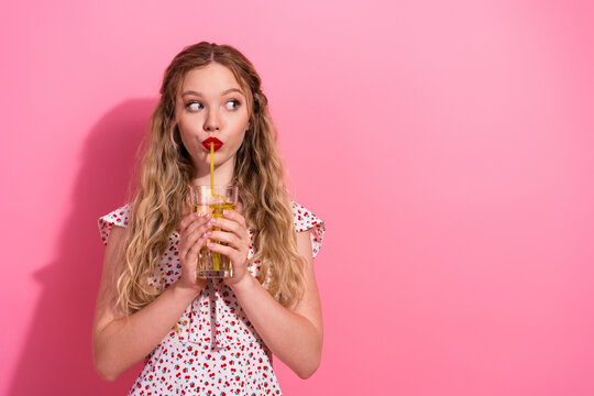 Young woman drinking juice through a straw against a pink background, playful portrait of a girl with red lips and wavy hair in a summer dress