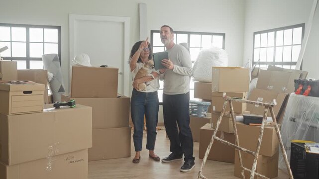 Middle age couple holding tablet and small dog amid stacked moving boxes inside building; moving day anticipation.