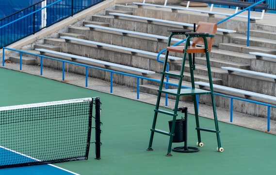 Empty outdoor tennis court featuring a prominent green umpire chair, black net, and concrete bleachers with blue railings in the background, ready for play or a championship match.