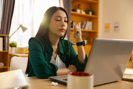 Stressed tired asian woman sitting at home looking at laptop screen feeling sick needing pill medicine for headache working at desk with exhaustion and anxiety during morning sunlight