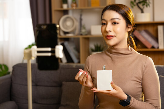 Happy young woman presenting perfume bottle to smartphone camera on tripod while recording video vlog at home in cozy living room setting