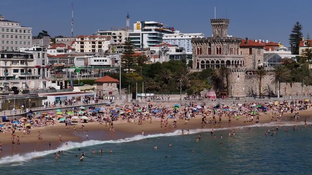 Estoril, Portugal - 18 April 2026: Aerial view of Tamariz Beach crowded with people sunbathing and swimming near the historic Forte da Cruz castle and city skyline.