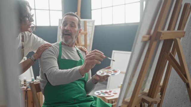 Man middle age paints at easel with palette and brush while woman leans in and rests hand on his shoulder in art studio; calm mentorship.