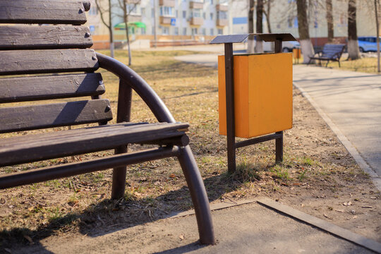 Yellow metal trash bin in a city park next to a brown wooden bench and a pedestrian path. Municipal metal trash container