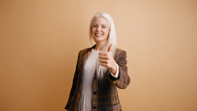 Woman smiling and giving a thumbs up gesture, showing approval, success, and positivity, standing against a neutral background
