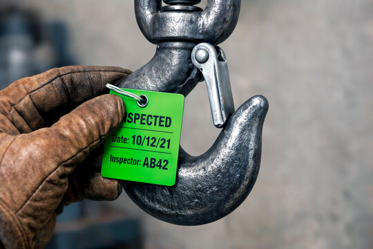 Gloved hand attaching green inspected tag to a metal lifting hook.
