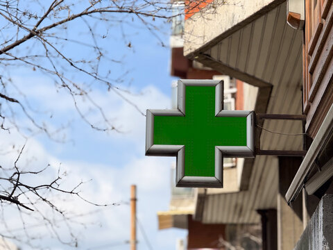 Green pharmacy cross sign mounted on building facade with sky and tree branches. Healthcare service symbol, medical assistance, drugstore identification and urban navigation marker.