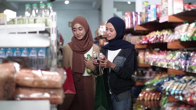 Asian muslim friends reading product label and comparing ingredients in supermarket aisle, one recommending beverage while other