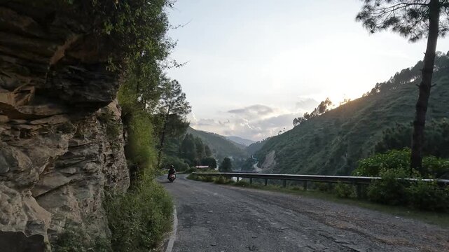 Point of view driving on a winding mountain road during sunset with scenic hills, trees and a scooter ahead. Beautiful natural landscape in India ideal for travel, adventure and road journey concepts.
