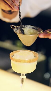 Bartender carefully straining a yellow cocktail through a fine mesh sieve into a glass with a large ice cube and sugar rimmed edge