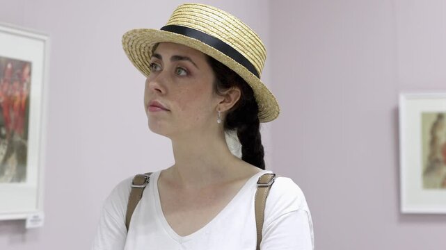 Pretty young elegant Caucasian woman wearing straw hat contemplating exposition with arts. Exhibition in gallery. Cultural education and Museum's Day