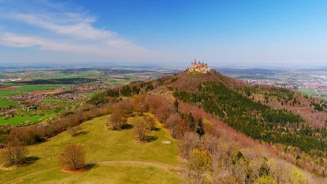 Burg Hohenzollern, Germany - 15 April 2026: Aerial view of Hohenzollern Castle perched on a hilltop surrounded by dense forests and rolling green fields under a clear blue sky.
