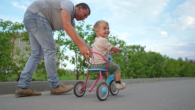 A happy father pushes his son on a tricycle in a green park. This cute little boy smiles while riding the tricycle outdoors. The loving father helps his son to ride. Happy family bonding is fun.
