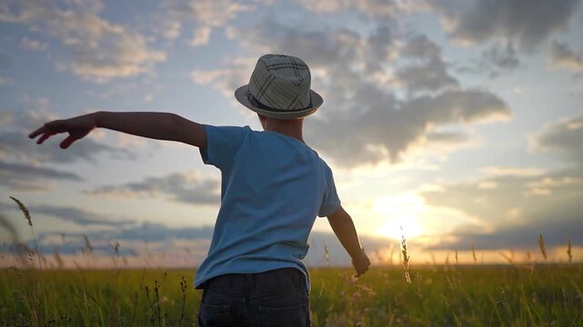 Boy stands in green field with arms outstretched at sunset. Happy child in hat plays pilot in summer nature. Kid dreams of flying with open arms in field. Boy enjoys freedom in sunny meadow.
