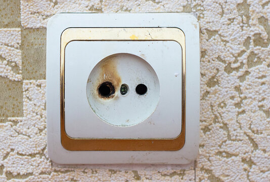 Dirty power outlet with burned hole on wallpapered wall. Short circuit hazard.