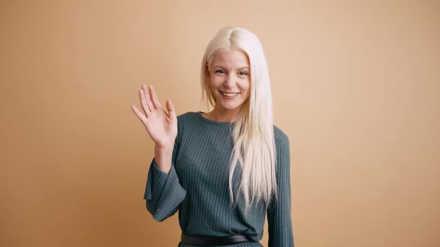 Cheerful smiling woman with long platinum blonde hair looking at the camera and waving her hand in a greeting gesture in a studio