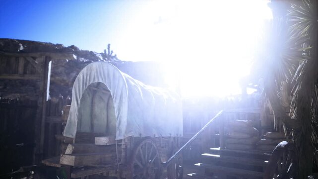 Sunlit pioneer wagon encampment with canvas covers and intense lens flare, wooden steps and rustic outbuilding bathed in blinding glare, deep dust ruts leading