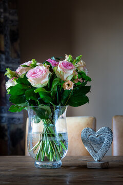 Eleganter Blumenstrau&szlig; mit rosa Rosen in Glasvase auf rustikalem Holztisch neben Deko-Herz aus Silber vor neutralem Hintergrund, romantisches Stillleben f&uuml;r Muttertag oder Liebe