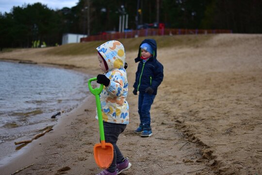 Two small children siblings playing on sandy lake shore with toys and sticks, outdoor activity in cool weather, childhood exploration and friendship in natural environment