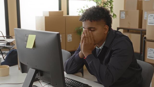 Man covering face with hands at computer amid parcel boxes in a building; stress overwork persistence.