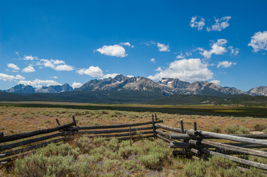Distant mountains rise above open grasslands. Weathered fence lines frame the scene. Blue sky holds scattered white clouds. Wild grasses blanket the foreground. Majestic peaks show rugged