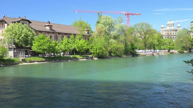 Sunny view across Aare river toward historic buildings, trees, and a construction crane in Bern, Switzerland.