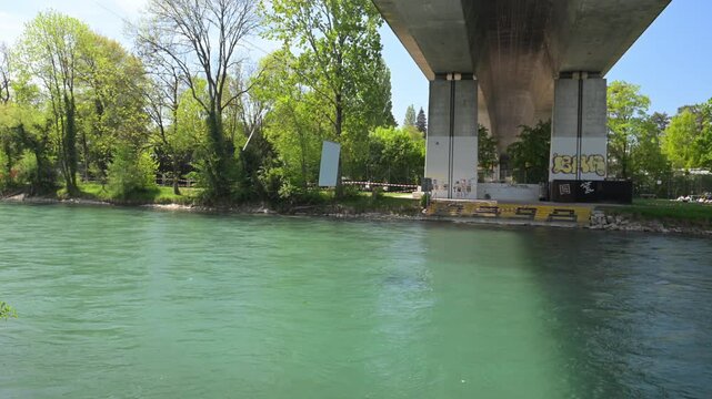 Bern, Switzerland - 26.04.2026: Concrete bridge spans turquoise river beside leafy park; graffiti visible on bridge supports.