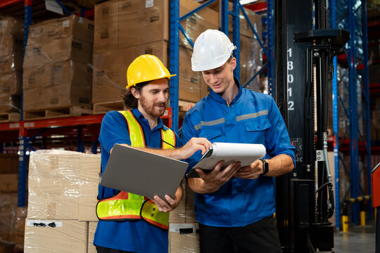 Male warehouse staff and supervisor reviewing logistics documents and shipment details together using clipboard and laptop. Professional collaboration in modern warehouse environment.