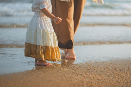 Asian toddler girl and parent walking barefoot on the beach during golden hour sunset. Concept of family travel, childhood memories, and summer vacation. A peaceful moment of a mother and daughter