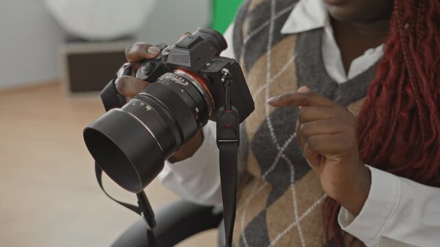 Woman points finger at camera while holding a mirrorless camera in a photo studio chair wearing argyle sweater vest and braided red hair; creative concentration.