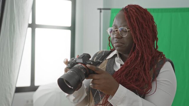 Woman photographer holding a dslr camera with hands, adjusting settings in a photo studio with green screen and softbox lighting; creative concentration.