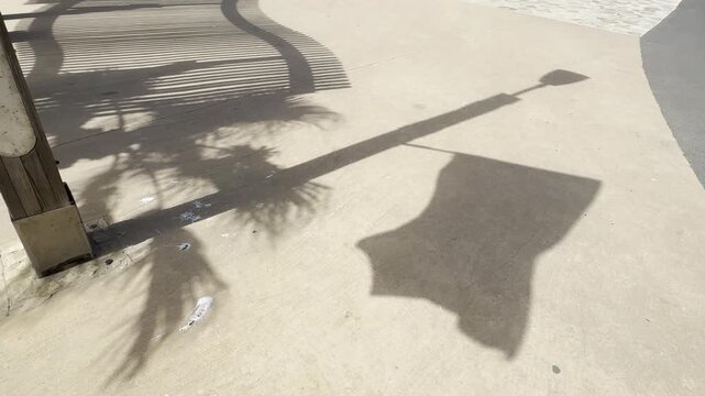 shadows of a flag and a palm tree moving on a sandy walkway next to a canopy shadow by the seaside