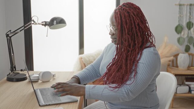 Woman types on laptop and adjusts glasses at a tidy home studio desk, hands on keyboard, lamp and headphones visible; focused productivity.
