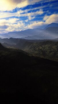 Hills and mountains are seen under a cloudy sky. The scene shows layers of hills fading into the background. Light shadows are cast on the land as clouds drift by.