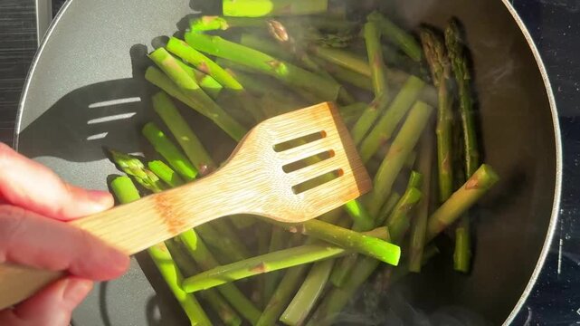 Top view of asparagus being stirred in frying pan. Healthy cooking, organic vegetables, food preparation, vegetarian meal, kitchen process, clean and minimal food concept.
