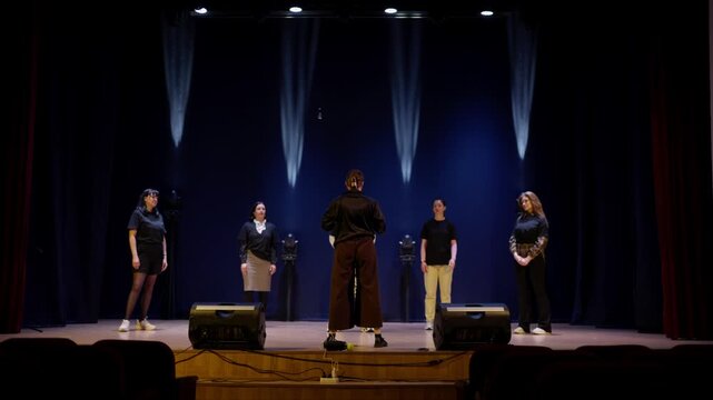 Stage rehearsal with vocal performers standing in formation under dramatic blue lighting as a central conductor leads a focused ensemble on an empty theater stage