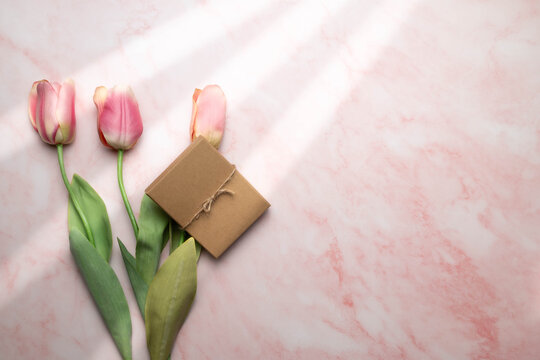 Sun rays shining on three pink tulips with a brown paper gift box on a pink and white marbled surface.  Mother's, woman's day, or birthday background.