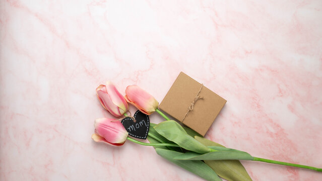 Three pink tulips with a brown paper gift box and heart shaped tag with Mom, on a pink and white marbled surface.  Mother's Day, or birthday banner.