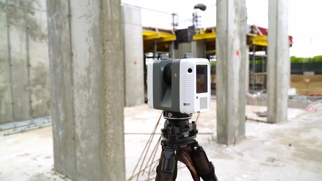 Device mounted on tripod at construction site. Large concrete pillars surround equipment. Background shows scaffolding and ongoing work. No people or animals visible in the scene